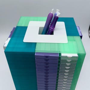 May include: A square storage organizer featuring translucent teal and light green panels, a stack of purple drawers, and a column of clear drawers. A white square top plate with a central opening holds two purple and teal gradient stylus tools.