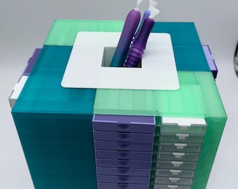 May include: A square storage organizer featuring translucent teal and light green panels with a central white square top plate. A vertical column of purple drawers sits beside a column of clear drawers. Two gradient purple pens are inserted in the center.