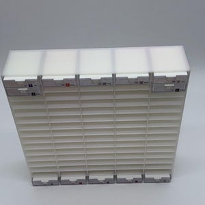 May include: A white, rectangular storage unit with multiple horizontal slots. The top section is solid, while the main body features a slatted design. Small labels with numbers and letters are visible on the front.