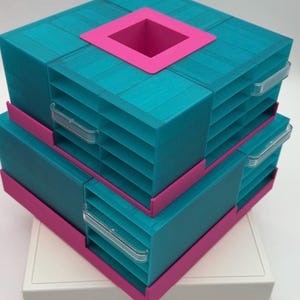 Puede incluir: Un organizador de almacenamiento modular con dos unidades apiladas de plástico color verde azulado con múltiples cajones y tiradores transparentes. La unidad superior tiene una abertura cuadrada rosa en el centro y bordes rosas sobre una base blanca.