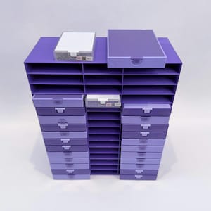 May include: A purple and lavender storage unit with multiple drawers and shelves. Two small white boxes with text are on top. The unit is designed for organizing and storing small items. The drawers have a pull-out design.