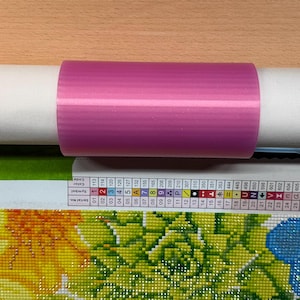 May include: A pink plastic roll holder is placed on a white paper tube. The tube rests above a partially visible diamond painting with a floral design. The painting features yellow, green, and blue colors.