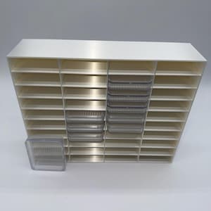 May include: A white storage unit with multiple compartments and horizontal shelves. Clear plastic trays are placed in some sections. The design is for organizing small items, with a clean, minimalist aesthetic. The unit is likely made of plastic.