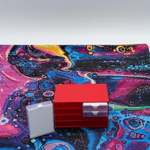 Puede incluir: Una unidad de almacenamiento de plástico rojo con múltiples ranuras horizontales y dos cajones transparentes, colocada junto a un pequeño recipiente rectangular de plástico blanco translúcido, sobre un lienzo de diamond painting texturizado.