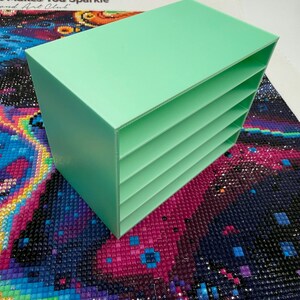 May include: A mint green rectangular storage organiser featuring four horizontal shelves stacked vertically. The unit has a solid back and side panels with open front access to each shelf, providing a clear view of the interior compartments.