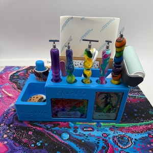 Peut inclure: Un support organisateur en plastique bleu marqué Sparkle et Brightly, contenant cinq stylos colorés à motifs avec des pointes en métal, un petit capuchon bleu, un objet en forme de biscuit et un rouleau bleu clair. Deux cartes sont insérées.
