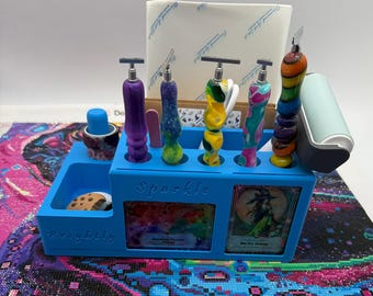 May include: A bright blue plastic organizer with the words Sparkle and Brightly embossed on the front. It holds five multi-colored diamond painting pens, a small blue wax container, and a light blue roller. Two rectangular art cards are inserted.