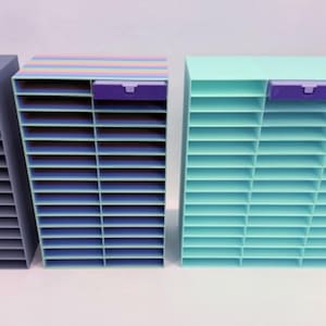 May include: Three colorful, tiered storage units. The units are in shades of gray, blue, and teal, with purple accents. Each unit has multiple horizontal shelves and a small, purple drawer at the top. These organizers are ideal for office or craft supplies.