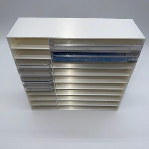 May include: A white plastic storage organiser featuring a vertical column of small clear drawers on the left and a series of wide horizontal shelves on the right. The top two shelves contain clear trays, with one tray holding small blue beads.