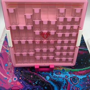 May include: A light pink plastic storage organiser with multiple clear rectangular compartments for small items. The front features a red diamond logo and the text Diamond Art Club DO WHAT MAKES YOU SPARKLE. It includes a matching pink stand.