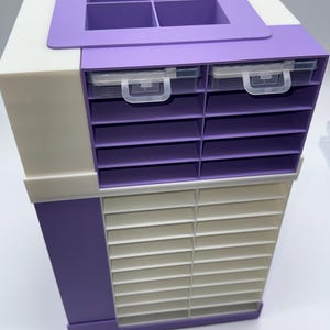 May include: A multi-tiered storage organizer featuring a white and purple frame. The top section has four square compartments, two clear plastic drawers with handles, and ten purple shelves. The bottom section contains twenty-four white slots.