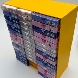 Pode incluir: Uma unidade de armazenamento amarela com várias gavetas pequenas em tons de azul, rosa e branco. Cada gaveta é rotulada com números e letras. A unidade foi projetada para guardar itens pequenos.