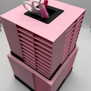 Puede incluir: Un organizador de almacenamiento cuadrado de color rosa de dos niveles con múltiples ranuras horizontales en cada lado. La parte superior tiene una abertura cuadrada negra central que contiene tijeras blancas y una herramienta rosa.