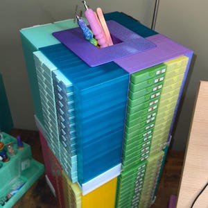 May include: A multi-colored, square, tiered storage tower featuring numerous small drawers in shades of teal, blue, green, yellow, and red. The top surface has a central square opening holding several craft tools, including a glittery blue pen and a pink tool.