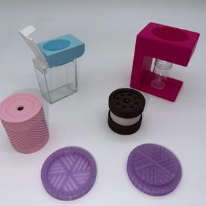 May include: A collection of 3D printed items including a light blue and clear plastic dispenser, a magenta stand with a clear vial, a textured light pink cylindrical container, a dark brown cookie-shaped object, and two purple circular textured coasters.
