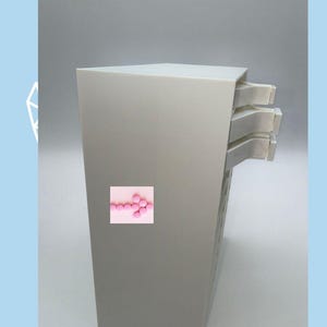 May include: A tall, light gray storage unit with three stacked drawers extending from the right side. A small square graphic featuring a pink arrow made of circular dots is on the front panel. The text 'Sale' and 'Item 2' appear in the image.