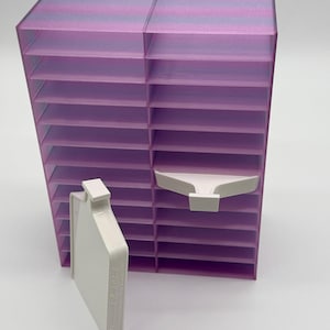 May include: A vertical storage tower featuring multiple horizontal shelves with a translucent purple and pink striped pattern. Two white plastic trays are included, with one tray positioned horizontally on a shelf and the other standing upright.