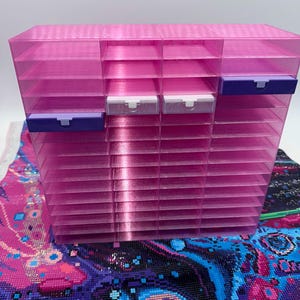 May include: A translucent pink plastic storage organiser featuring multiple horizontal slots for small items. The unit includes two dark purple drawers on the left and right sides, and two clear white drawers positioned in the centre rows.