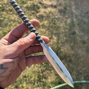 以下が含まれることがあります： 手作りの金属製のナイフ。 質感のあるハンドルと、研磨された鋭利な刃が付いています。 ハンドルには一連のリッジがあり、刃は銀色です。 ナイフは、緑の草と日光を背景に保持されています。