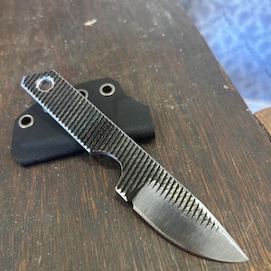 May include: A small, silver knife with a textured black handle and a black sheath. The knife blade is sharp and pointed. The handle has a hole for a lanyard. The knife is resting on a dark wooden surface.