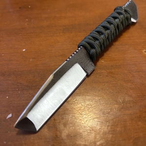 Könnte beinhalten: Ein kleines Messer mit silberner Klinge und einem dunkelgrünen, gewebten Griff. Die Klinge hat eine scharfe Kante und eine strukturierte Oberfläche. Der Griff ist eng mit einer dunkelgrünen Kordel umwickelt.