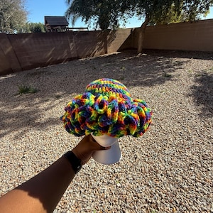 May include: A colorful, rainbow-hued crochet hat with a wide brim. The hat is displayed on a white mannequin head. The hat's design features a vibrant mix of colors, including red, orange, yellow, green, blue, and purple.
