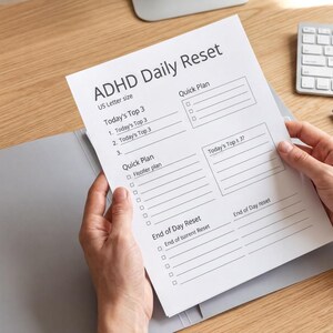 A person's hands are holding a sheet titled "ADHD Daily Reset" on a wooden desk, with a keyboard and a coffee cup visible in the background.