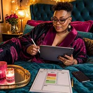 A woman in a purple robe is sitting on a bed, using a tablet and surrounded by various items, including a candle, a tray, and a notebook.