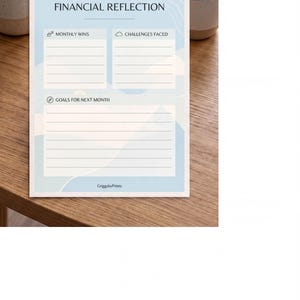 a financial reflection form with blank spaces for recording monthly wins and challenges faced. The form is placed on a wooden table, with a potted plant and a cup visible in the background.