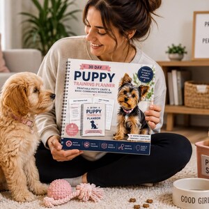 A woman sits on the floor holding a "30 Day Puppy Training Planner" book, surrounded by two small dogs and various dog toys.