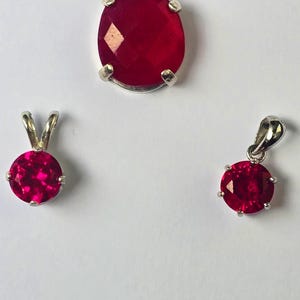 May include: Three silver pendants with vibrant red gemstones. The largest pendant has an oval-shaped stone, while the other two feature round stones. The gemstones are set in silver settings. The image is on a white background.