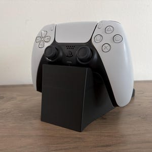 以下が含まれることがあります： 黒い充電スタンドに置かれた、白と黒のPlayStation 5コントローラー。 コントローラーには、タッチパッド、ボタン、アナログスティックがあります。 スタンドは角度がついています。