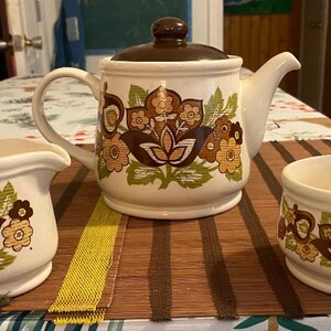 May include: A vintage cream-colored tea set with a teapot, creamer, and sugar bowl. Each piece features a brown and orange floral design with green accents. The teapot has a brown lid and a curved spout.