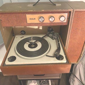 Könnte beinhalten: Vintage RCA Plattenspieler mit hellbraunem Holzgehäuse und Lautsprecher auf der rechten Seite. Der Plattenteller ist weiß mit einer schwarzen Schallplatte und einem schwarzen Tonarm. Das Bedienfeld hat drei Knöpfe und das RCA-Logo.