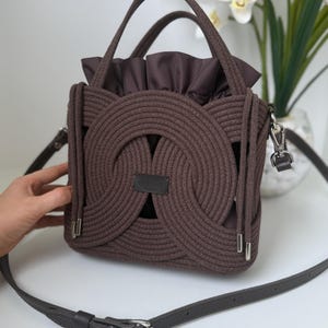 Könnte beinhalten: Braune Handtasche im Seil-Stil mit quadratischer Form und einem dunkelbraunen Lederriemen. Die Tasche hat ein einzigartiges Webdesign auf der Vorder- und den Seiten, mit einem gerafften Stoffinnenraum. Sie hat zwei Griffe und einen abnehmbaren Schulterriemen.