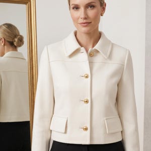 Puede incluir: Una chaqueta corta de color crema con cuello estructurado, mangas largas y cuatro botones circulares dorados en la parte delantera. La chaqueta incluye dos bolsillos rectangulares con solapa en la cintura y una costura horizontal en el pecho.