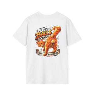 May include: A white t-shirt featuring a graphic of an orange tabby cat with its back turned. The text reads "It Takes BALLS to TNR" above the cat and "TRAP NEUTER RETURN" on a banner below, with the cat's tail raised and visible anatomy.