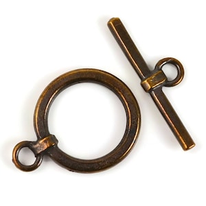 May include: A bronze-colored toggle clasp, a jewelry finding, lies on a white background. The clasp consists of a circular ring with a small loop and a straight bar with a loop at one end. The metal has a slightly aged appearance.