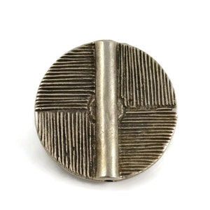 May include: A circular silver-toned metal button featuring a raised vertical bar across the center. The surface is divided into four quadrants, each decorated with a pattern of fine, parallel etched lines running in alternating directions.