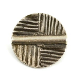 May include: A circular silver-toned metal button featuring a raised horizontal bar across the center. The surface is divided into four quadrants, each decorated with a pattern of fine, parallel etched lines running in alternating directions.
