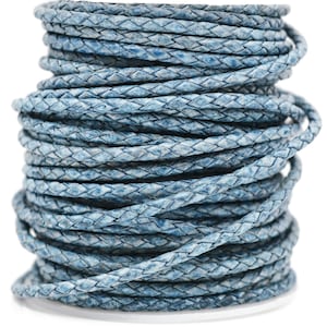 May include: A spool of braided cord in a dusty blue color with a textured, weathered appearance. The cord is wound into a tall, cylindrical stack, showcasing the intricate woven pattern of the material throughout the entire length of the cord.