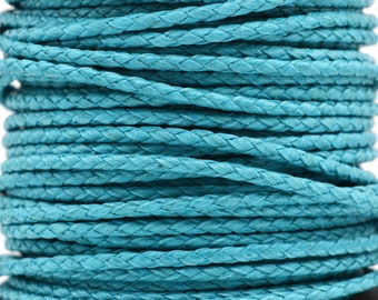 Puede incluir: Una bobina de cordón trenzado de color turquesa enrollado en múltiples capas. El cordón presenta una textura tejida consistente y un color azul verdoso vibrante y sólido a lo largo de toda la longitud del material enrollado en la bobina negra.