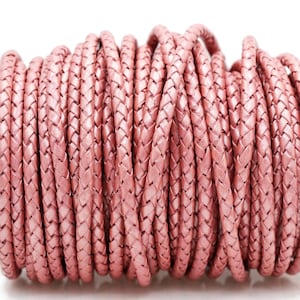 May include: A spool of braided, rose-colored leather cord. The cord is tightly woven, creating a textured surface. The roll is tightly wound, with the end of the cord visible. The background is plain white.