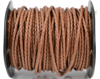 Puede incluir: Un carrete de cordón trenzado marrón enrollado alrededor de una bobina de plástico negro. El cordón presenta una textura tejida consistente en toda su longitud, mostrando un color marrón medio con variaciones sutiles en los hilos trenzados.