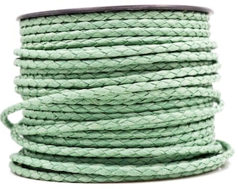 Puede incluir: Un carrete de cordón trenzado en color verde salvia claro. El cordón está enrollado firmemente alrededor de una bobina de plástico negro, creando una forma cilíndrica con una textura tejida visible a lo largo de todo el material, mostrando un trenzado consistente.