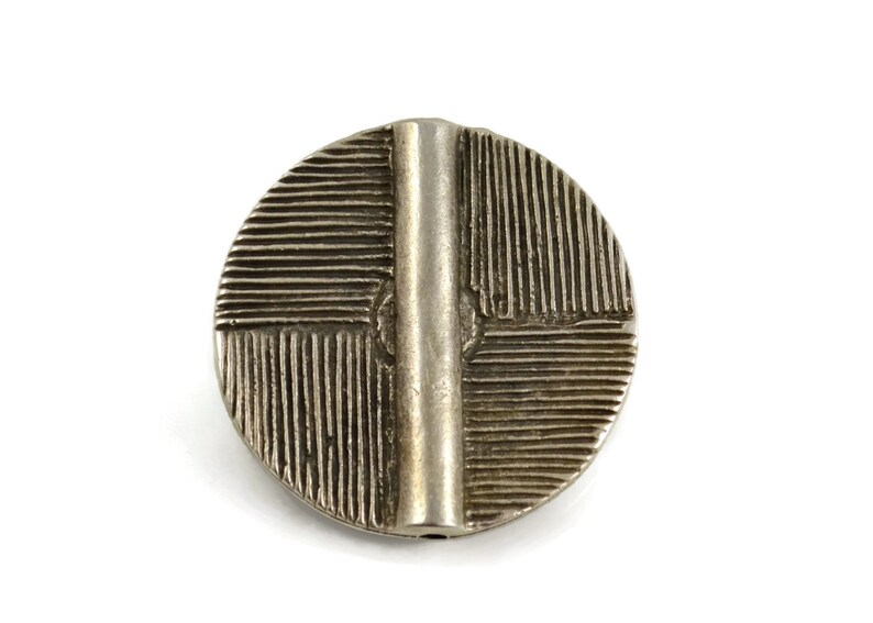 May include: A circular silver-toned metal button featuring a raised vertical bar across the center. The surface is divided into four quadrants, each decorated with a pattern of fine, parallel etched lines running in alternating directions.