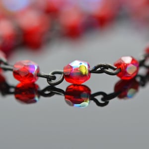 May include: A necklace featuring faceted, translucent red crystal beads with an iridescent finish. The beads are connected by dark, matte-finished metal links, creating a chain that reflects light and displays a vibrant, deep red color throughout.