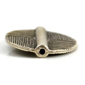 May include: A silver-toned metal bead with an oval, flattened shape and a central raised ridge. The surface features fine, etched line detailing radiating outward from the center. A single circular hole is visible at the end of the ridge.