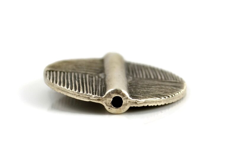 May include: A silver-toned metal bead with an oval, flattened shape and a central raised ridge. The surface features fine, etched line detailing radiating outward from the center. A single circular hole is visible at the end of the ridge.