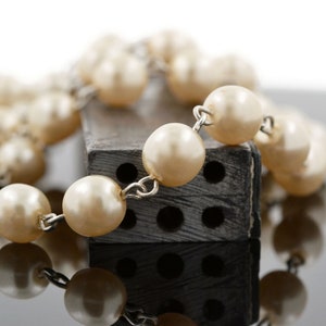 May include: A string of faux pearls draped over a black rectangular object with holes. The pearls are cream coloured and the object has a rough texture.